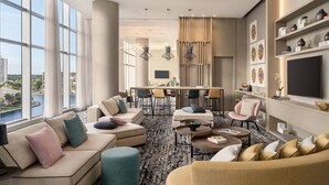 A modern hotel room with a large window offering a city view, a cozy seating area with sofas and ottomans, a dining area with a table and chairs, and a wall-mounted TV. - 2BR Oceanview HYDE – Free Parking, No Resort Fee (Hollywood)