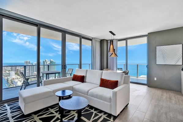 A modern living room with a large sofa, a small round table, and a view of the city and ocean through floor-to-ceiling windows.