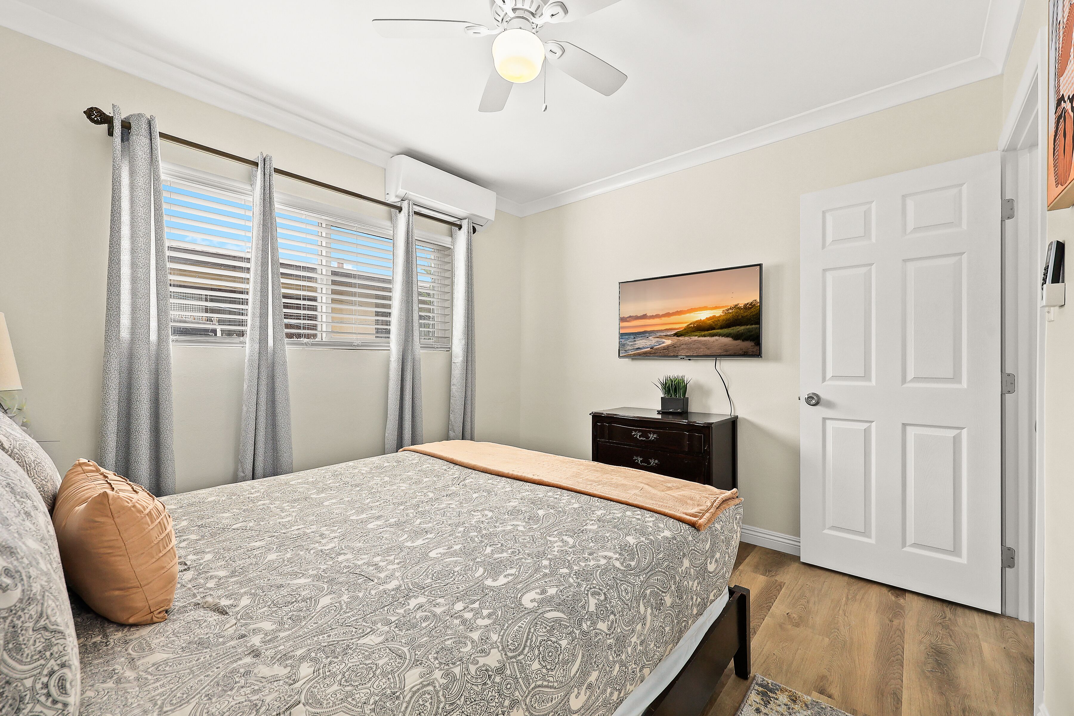 A bedroom with a bed, curtains, a ceiling fan, a TV, a dresser, and a door.