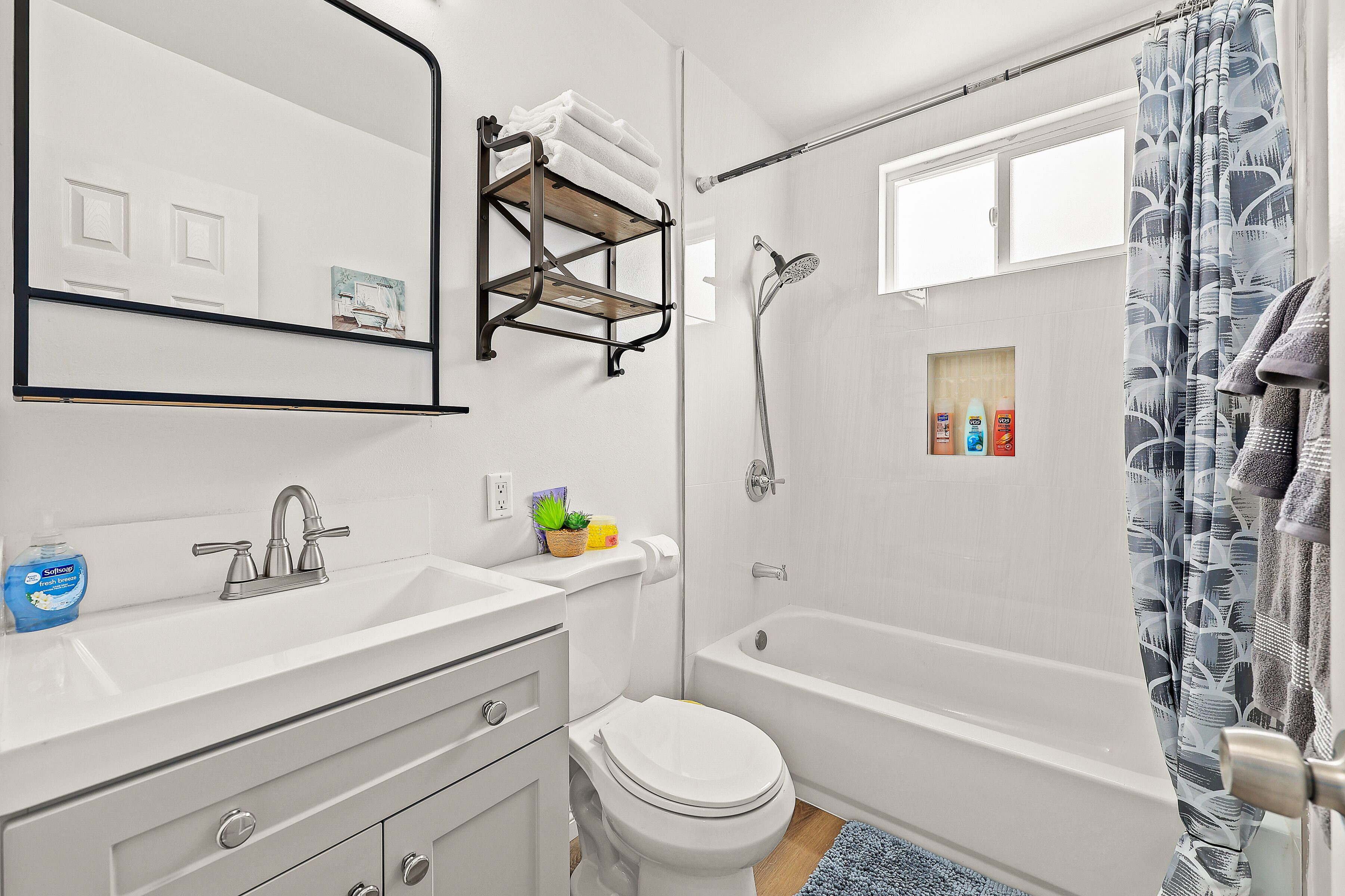 Et moderne badeværelse med en hvid vask, toilet og badekar. Der er et håndklædeholder over vasken og en brusegardiner med et mønster.