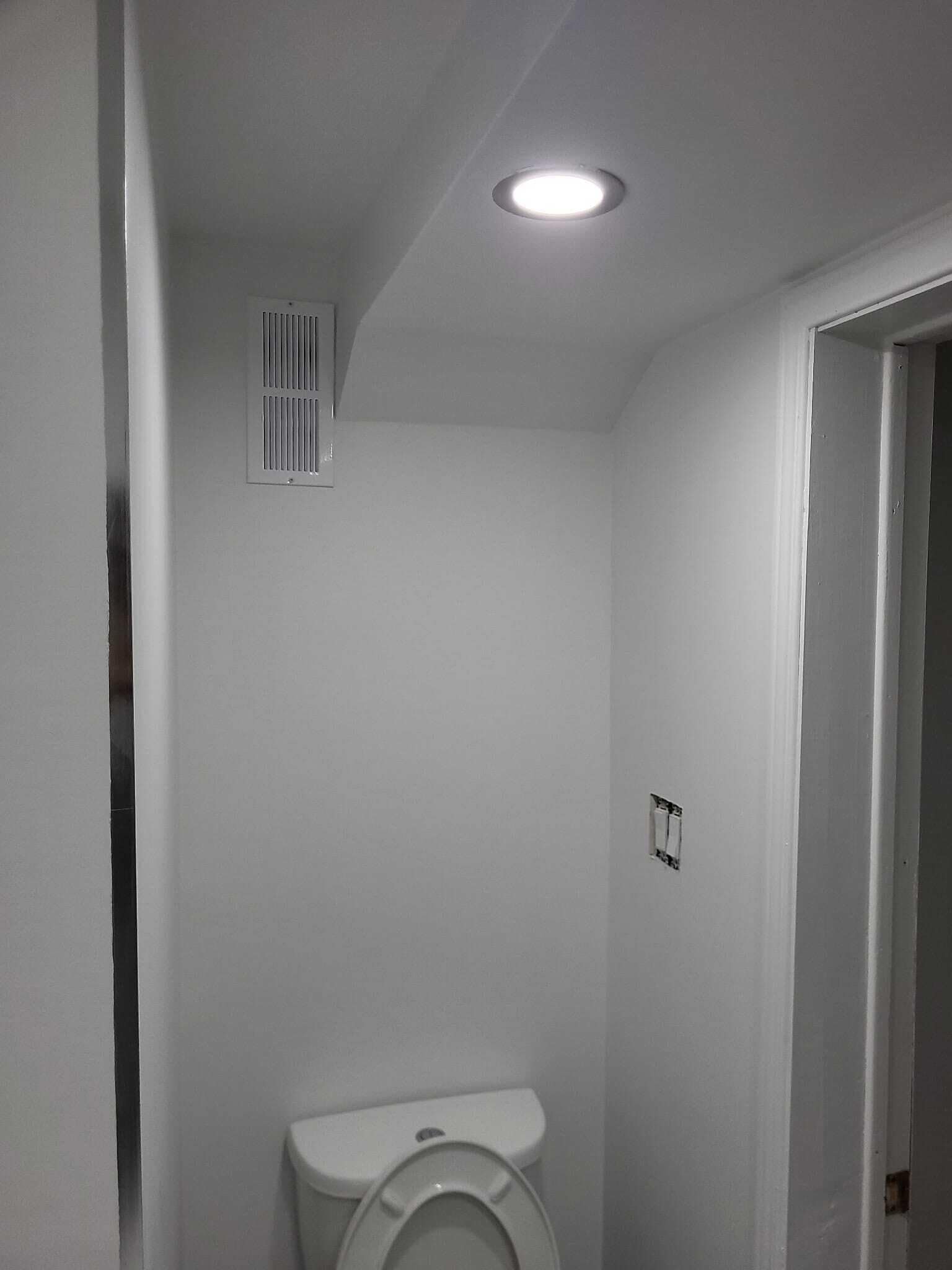 Uma casa de banho pequena e branca, com sanita, ventilação no teto e porta que dá para outro espaço.