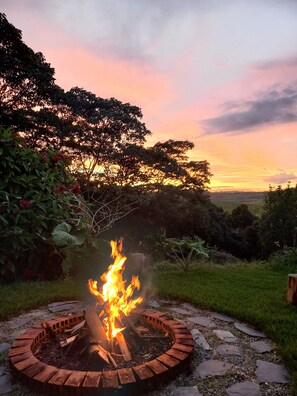 A circular fire pit with logs burning, surrounded by a stone border, set in a grassy area with trees and a sunset in the background. - Beautiful & Comfortable Suite In The Country Side (Circasia)