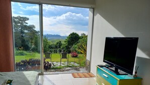 Une chambre avec une grande fenêtre, un lit, un téléviseur posé sur un meuble, et une vue sur un jardin et des collines.