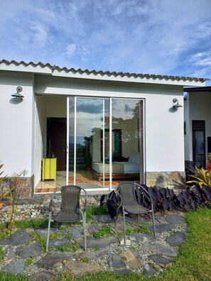 A modern house with a patio area featuring two wicker chairs, a yellow cabinet, and a glass door leading to an interior with a bed and a sofa. - Beautiful & Comfortable Suite In The Country Side (Circasia)