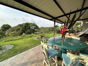 A covered patio area with a round table and chairs, surrounded by a garden with various plants and a view of the landscape. - Beautiful & Comfortable Suite In The Country Side (Circasia)