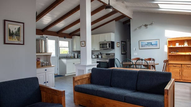 Ein gemütliches Wohnzimmer mit einem dunkelblauen Sofa und Holzstühlen, eine Küche mit weißen Schränken und modernen Geräten sowie ein Essbereich mit einem Holztisch und Stühlen.