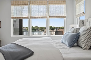 Une chambre avec un grand lit, donnant sur un balcon, et une fenêtre offrant une vue sur un immeuble et des arbres.