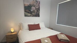 Una cama bien hecha con una colcha roja y blanca, dos toallas dobladas, una mesita de noche con una lámpara y un cuadro en la pared.