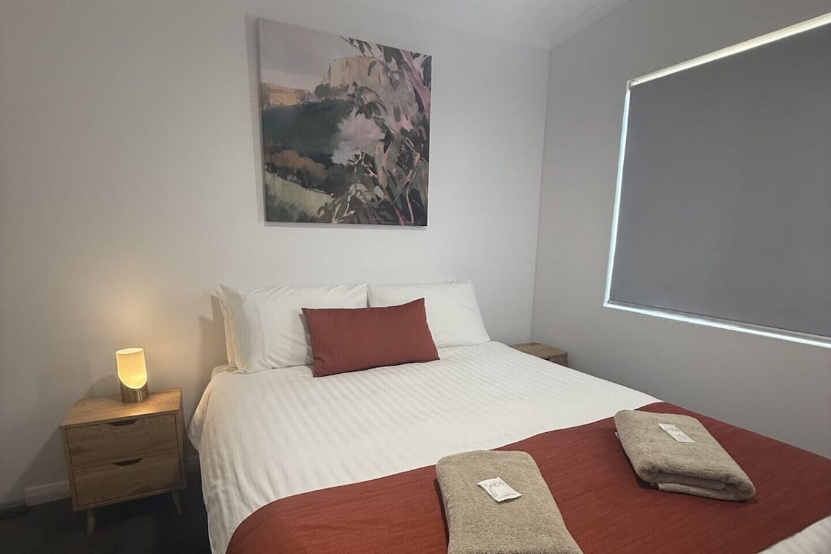 A neatly made bed with a red and white bedspread, two folded towels, a nightstand with a lamp, and a painting on the wall.
