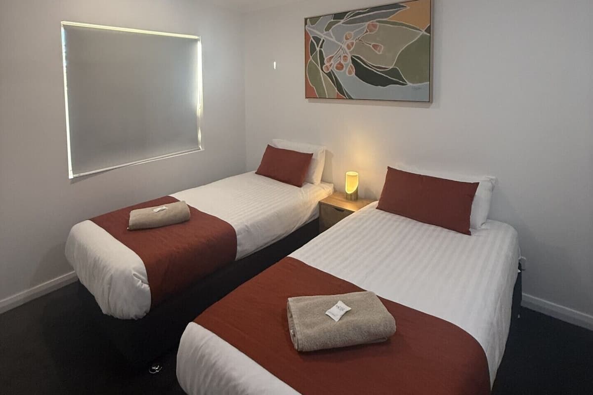 A hotel room with two beds, a ceiling fan, a window with blinds, a wall-mounted artwork, and a bedside lamp.