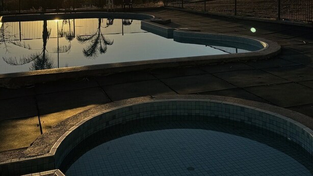 Una piscina al atardecer con reflejos nítidos, rodeada de una valla y palmeras.