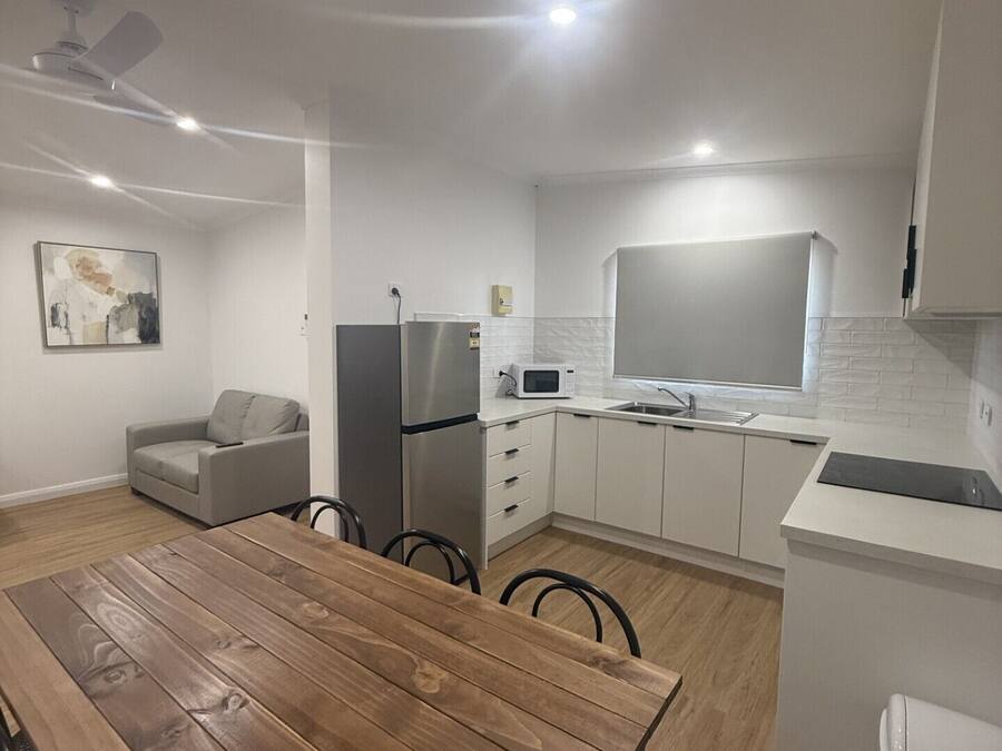 Eine kompakte Küche mit weißen Schränken, einem Edelstahlkühlschrank, einer Mikrowelle und einem Spülbecken. Es gibt einen Holzespeisetisch mit Stühlen, ein graues Sofa und einen Deckenventilator.