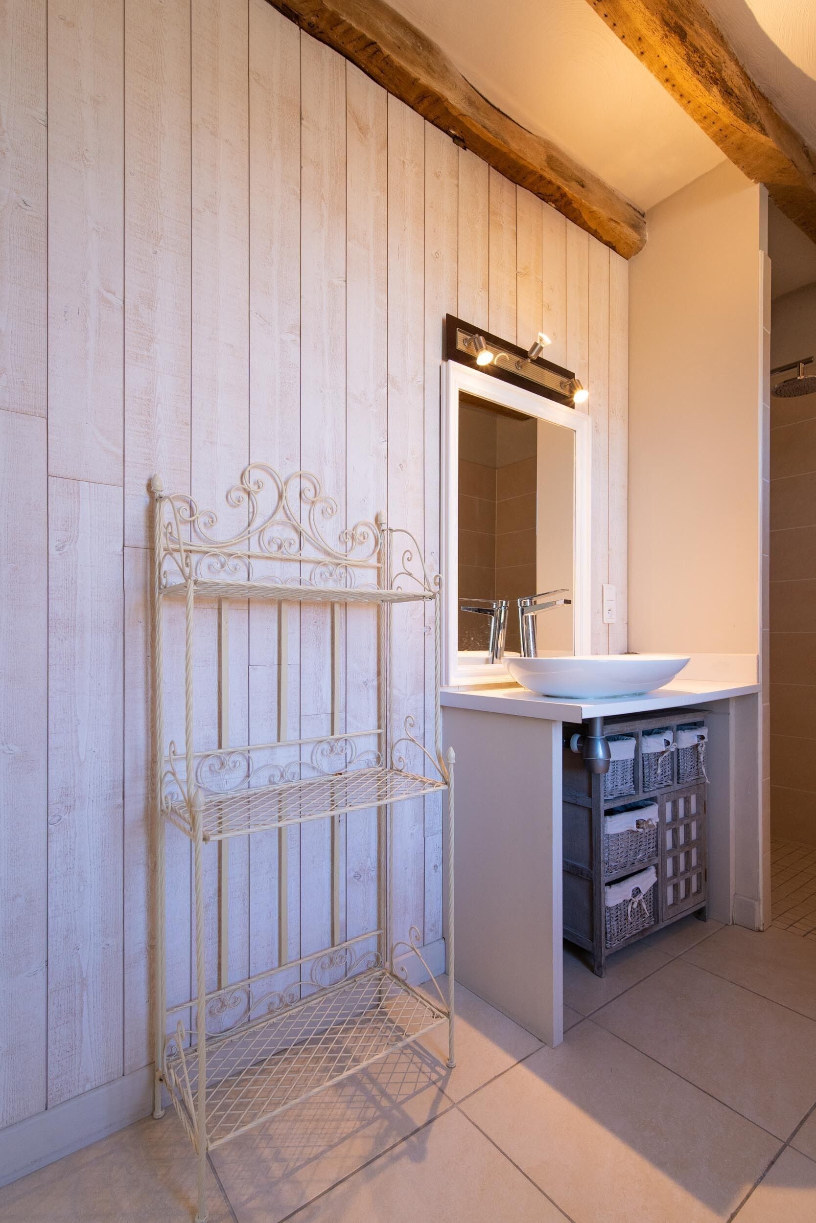 A bathroom with a white vanity, a mirror with lights, a metal rack, and tiled flooring.