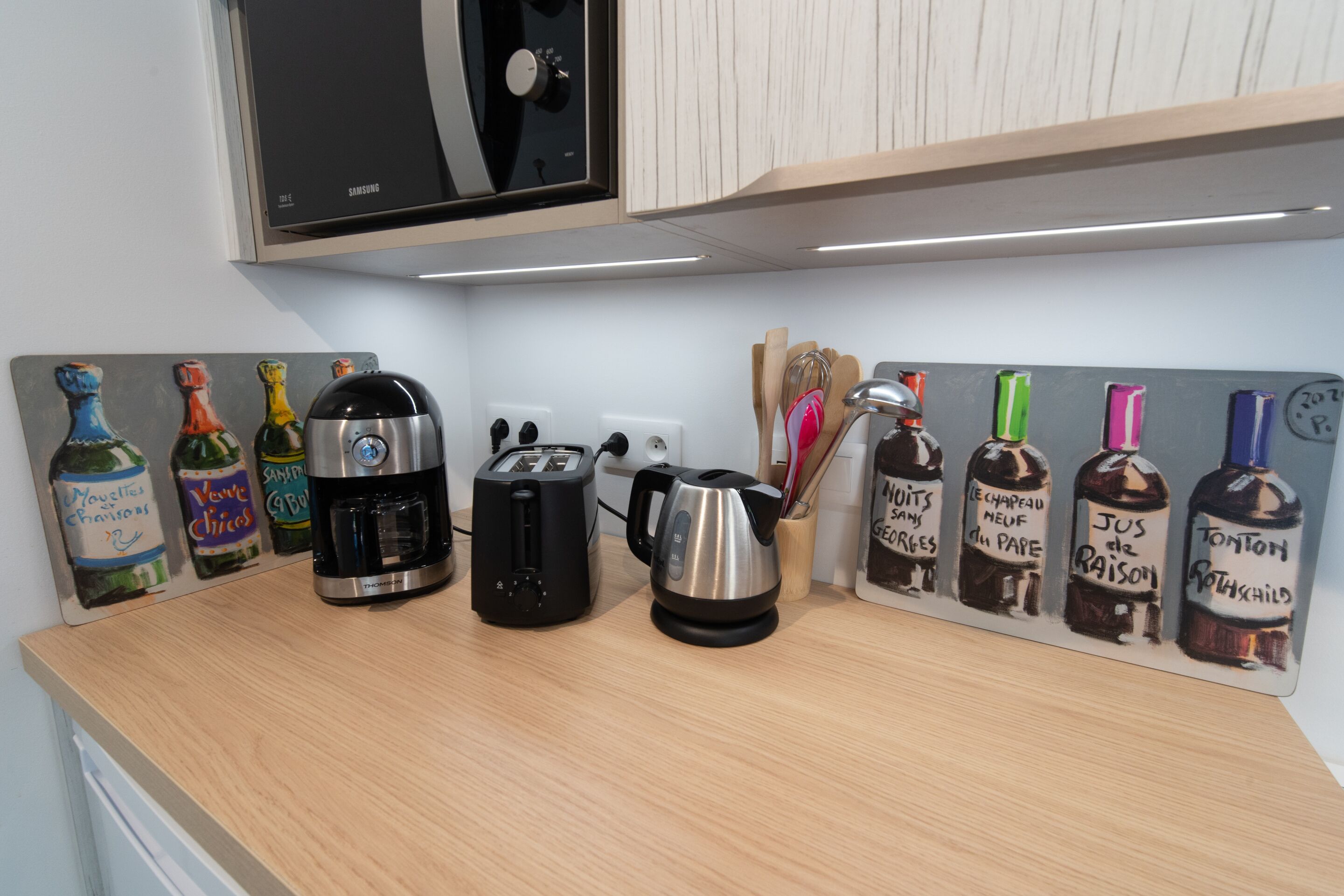 Een aanrecht met een broodrooster, waterkoker en koffiezetapparaat. Er staan ook wijnflessen op een muur.