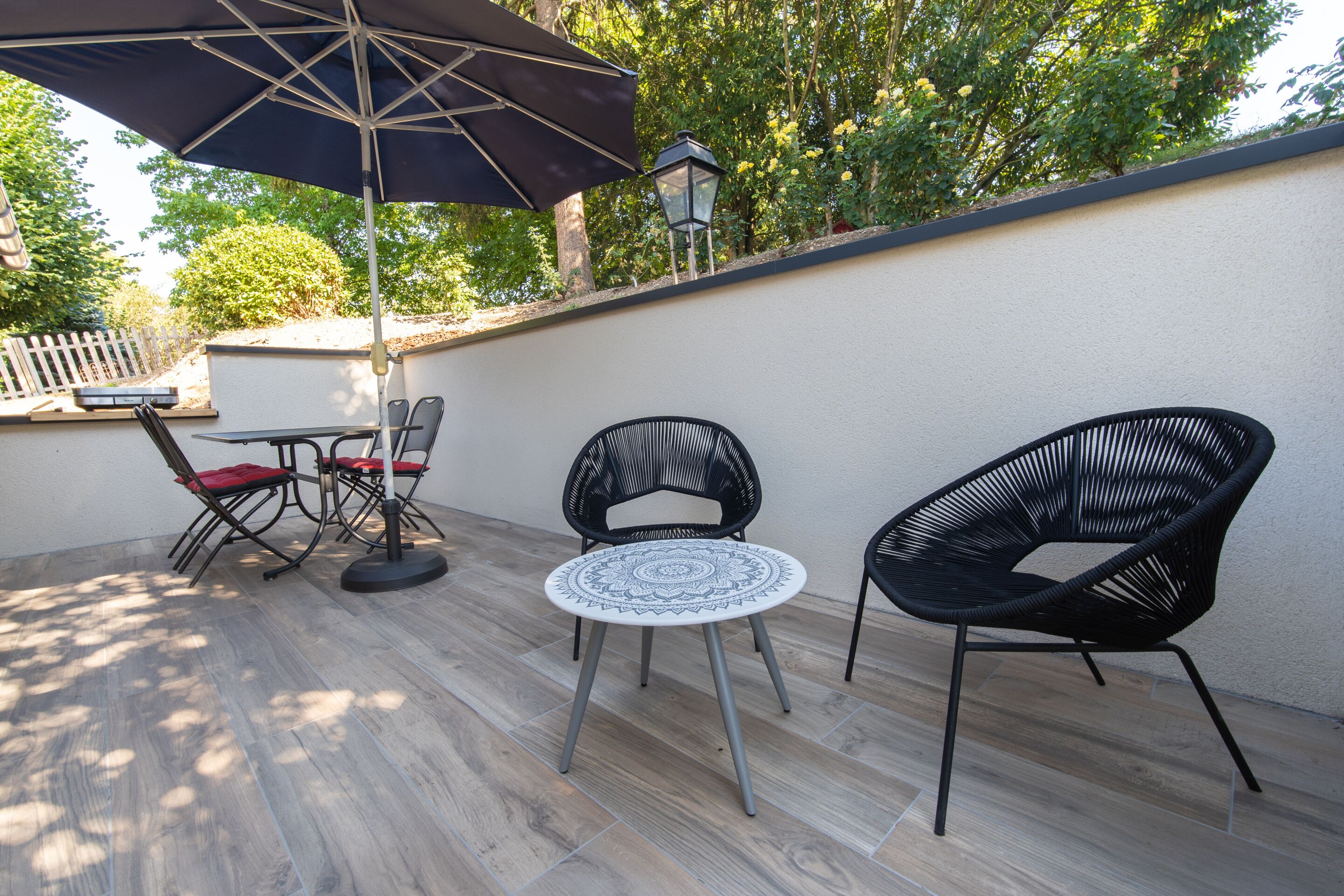 Een terras buiten met een tafel, stoelen, een grote parasol en een wandlamp.