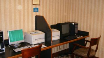 Habitación de hotel con un escritorio equipado con computadora de escritorio, una impresora y un televisor. Hay dos sillas de madera y una alfombra estampada.