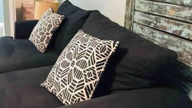 Ein dunkelbraunes Sofa mit zwei gemusterten Kissen, einer Lampe und einer rustikalen Holzwand.