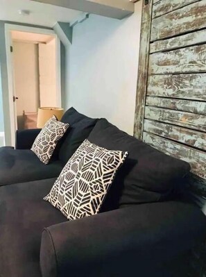 En mørk sofa med to puder med mønster, en lampe og en rustik trævæg.