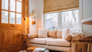 Een gezellige woonkamer met een houten deur, een beige bank, een vloerlamp en een groot raam met uitzicht op bomen.