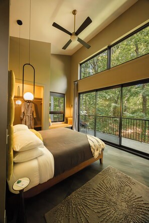 A modern bedroom with a large bed, a ceiling fan, and a view of the forest through large windows.