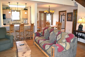 A living room with a sofa, chairs, a coffee table, and a dining area with a table and chairs. There is a kitchen in the background with cabinets and a sink.