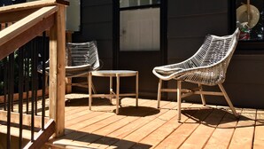 A wooden deck with two wicker chairs and a small table, a wooden railing, and a dark building facade with windows. - Parkman Pad: hot tub, wood stove by Montague breweries (Montague)