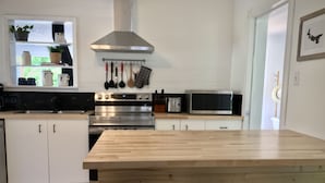 A modern kitchen with white cabinets, a wooden island with stools, stainless steel appliances, and a backsplash with hanging utensils. - Parkman Pad: hot tub, wood stove by Montague breweries (Montague)