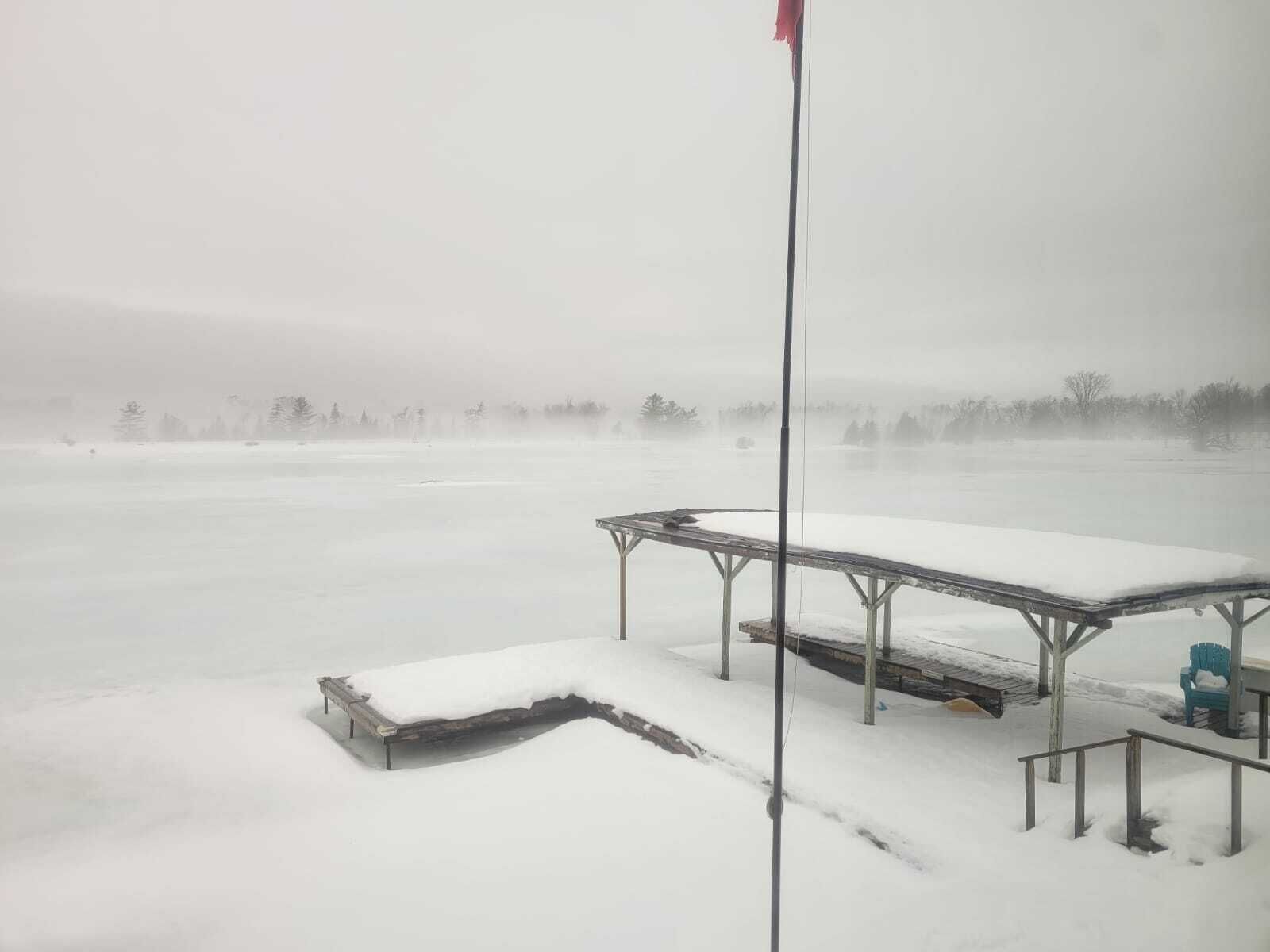A snow-covered outdoor area with a metal table and benches, a flagpole, and a distant view of trees and a body of water.