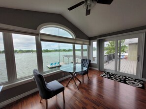 Una habitación con un gran ventanal con vista a un lago, una mesa de vidrio, dos sillas y un ventilador de techo.