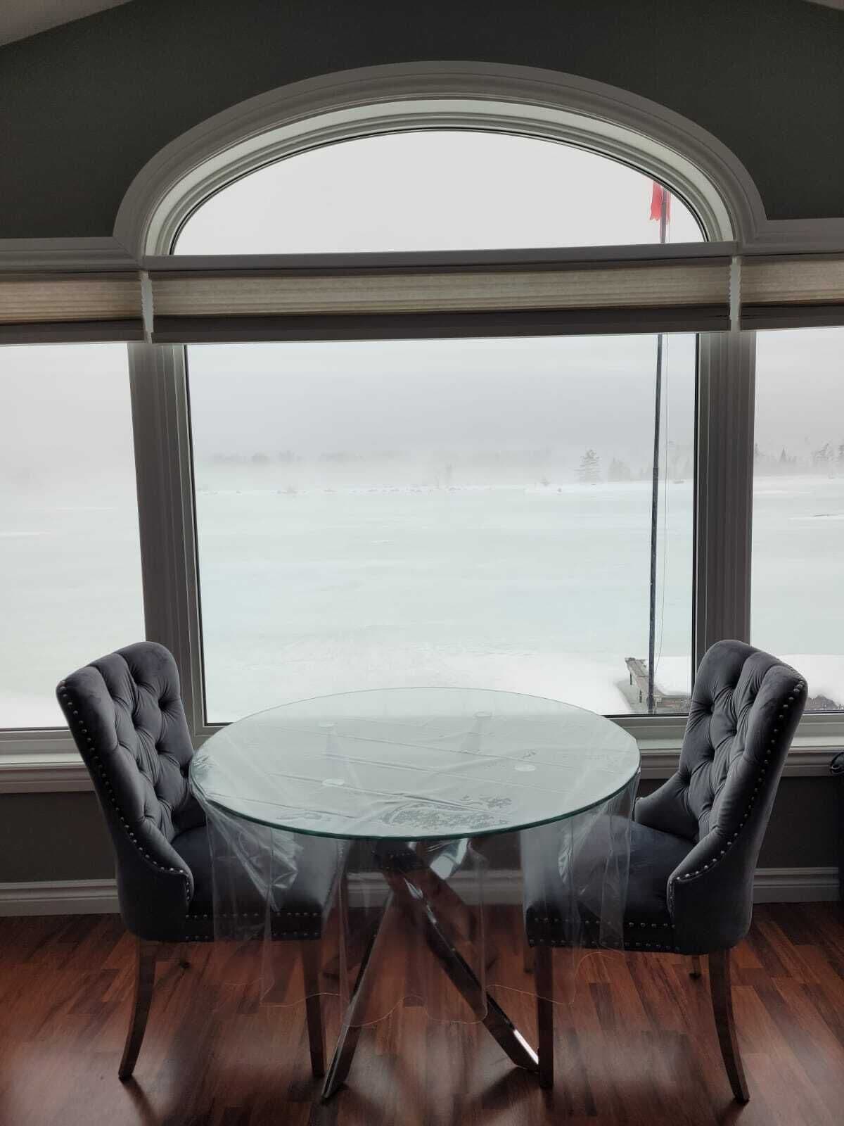 A room with a round glass table and two upholstered chairs, large windows with a view of a snowy landscape.