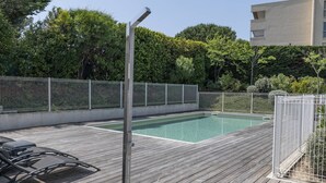 A swimming pool with a wooden deck, surrounded by a metal fence and lush greenery. - Superb 3P in a quiet location Swimming pool Terrace Barbecue Pétanque aircon (ANTIBES)