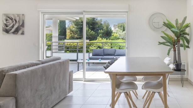 A modern living room with a grey sofa, a wooden dining table with chairs, a large potted plant, and a sliding glass door leading to an outdoor seating area with a grey sofa and a coffee table. - Superb 3P in a quiet location Swimming pool Terrace Barbecue Pétanque aircon (ANTIBES)