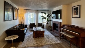 Een woonkamer met een donkerbruine leren bank, een houten salontafel, een klein rond tafeltje, een piano en een grote potplant.