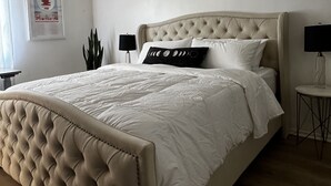 Een slaapkamer met een gestoffeerd hoofdeinde, een groot bed met wit beddengoed, twee nachtkastjes met lampen, een ingelijste poster aan de muur en een potplant.