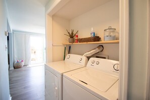 A laundry room with two front-loading washers, a shelf with a potted plant, cleaning supplies, and a basket.