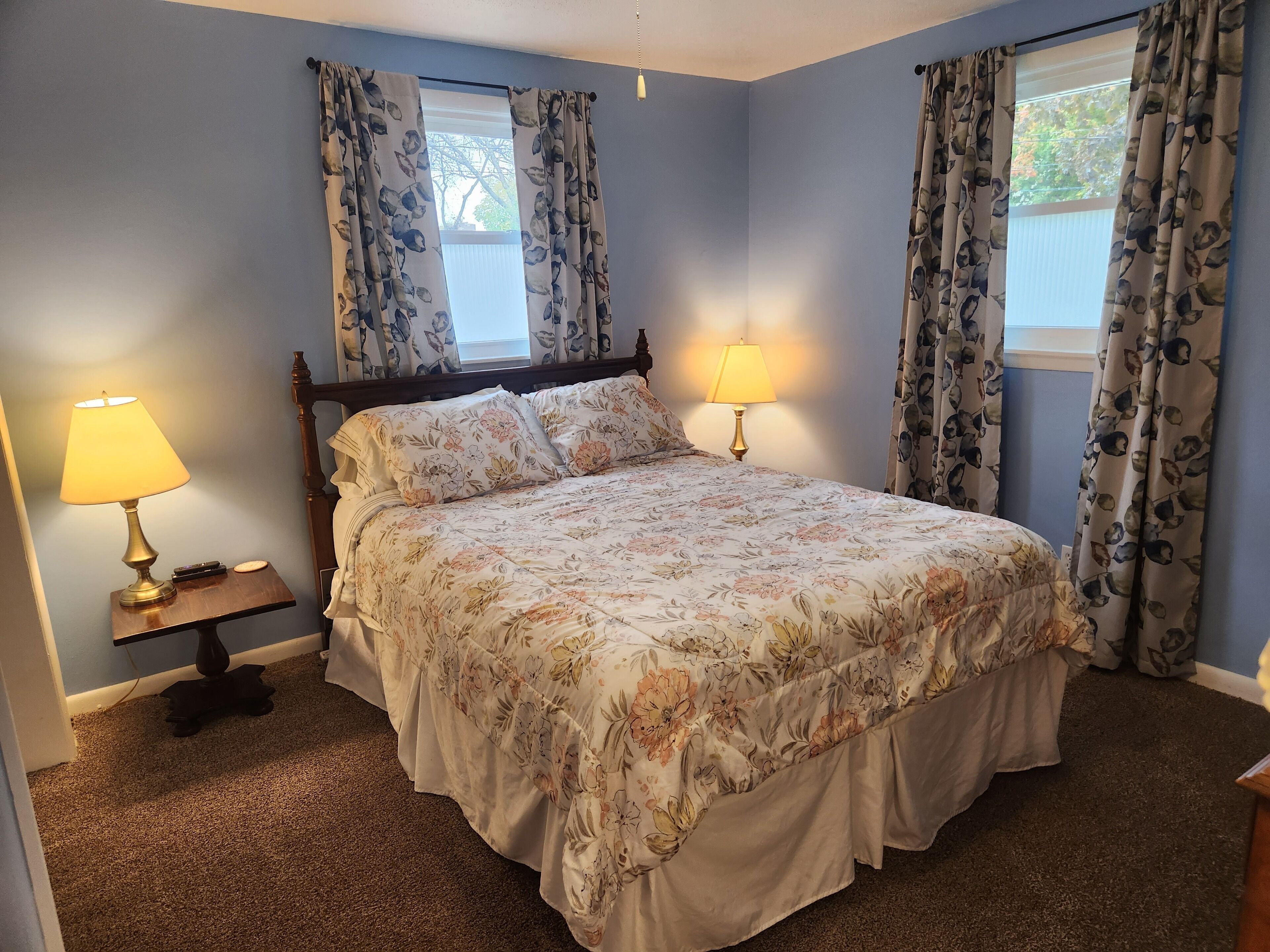 Une chambre à coucher avec une couvre-lit à motifs floraux, deux lampes de chevet et des rideaux aux deux fenêtres.