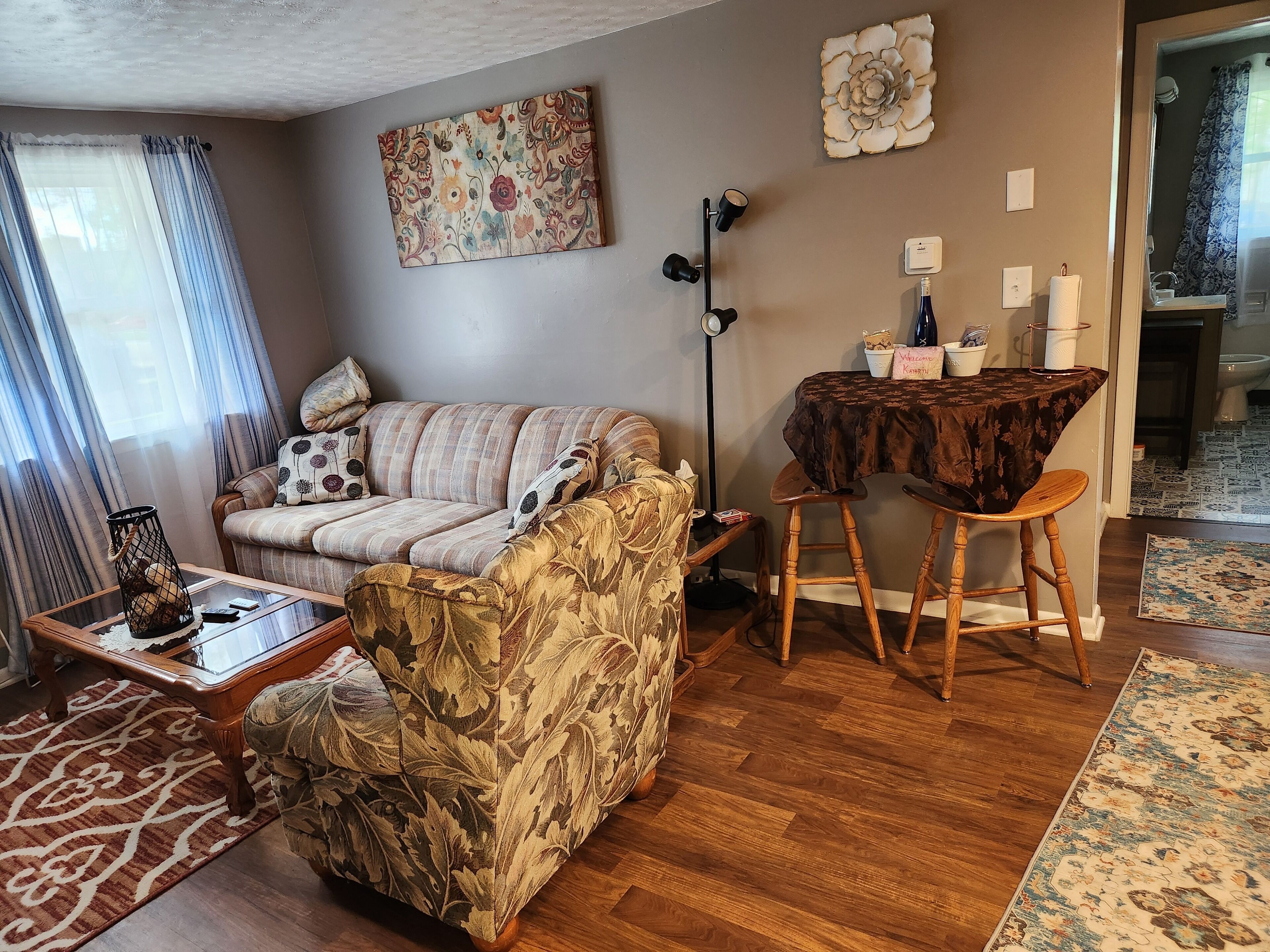 A living room with a sofa, a coffee table, a patterned armchair, a dining table with a chair, and a wall-mounted flower decoration.