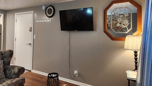 A living room with a flat-screen TV mounted on the wall, a clock, a coat rack, a patterned lamp, and a floral mirror.