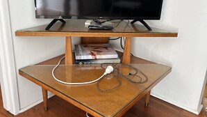 Un téléviseur posé sur un meuble en bois, avec une télécommande, des livres et un câble d’alimentation sur une table en verre.