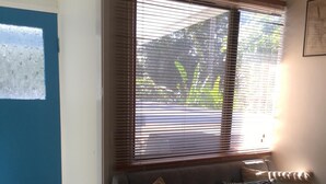Un salon avec un canapé gris, deux coussins, une fenêtre avec des stores, une porte et un sol carrelé.