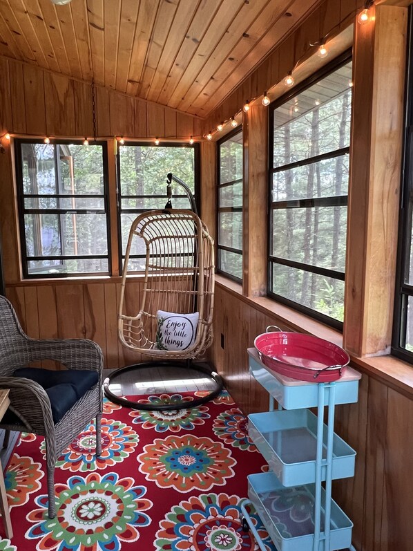 Une pièce chaleureuse avec un plafond en bois, de grandes fenêtres, un fauteuil en osier, un tapis coloré et une étagère bleue sur laquelle repose un récipient rouge.