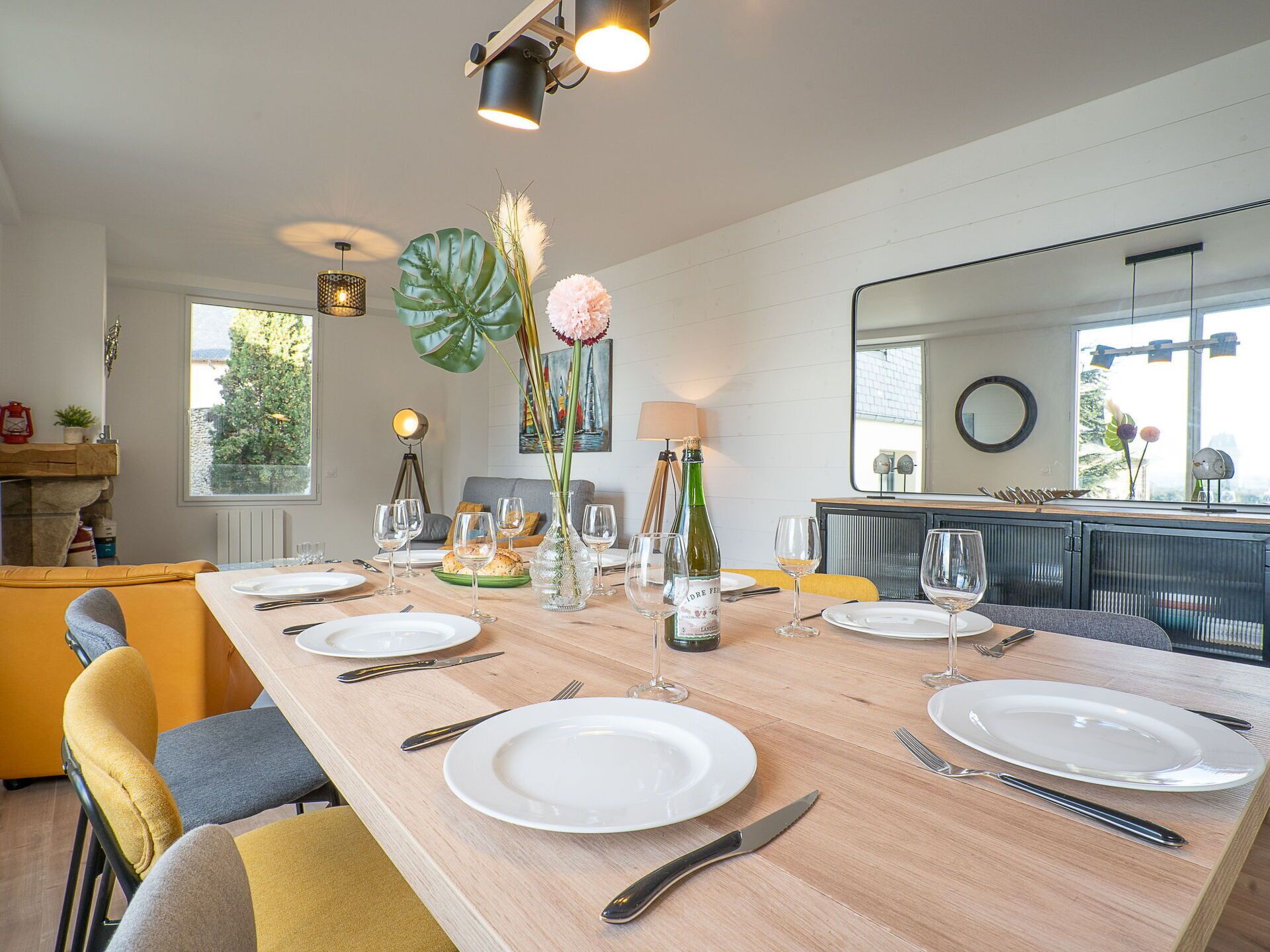 A dining area with a long wooden table set for a meal, a large window with a view, and a modern kitchen in the background.