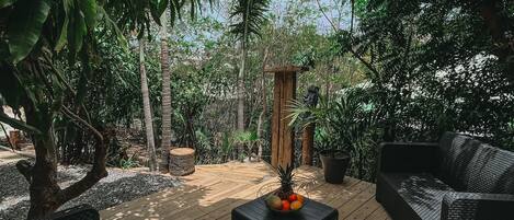 An outdoor patio with wicker furniture, a wooden table with fruit, and a pergola providing shade.