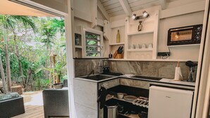 A compact kitchen with a built-in oven, a small refrigerator, and a window overlooking a garden. - Mango Cottage Orient Bay, Charming and Authentic (Collectivité de Saint-Martin)
