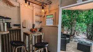 A cozy kitchen area with wooden stools, a bar counter, and a view of a lush garden through a window. - Mango Cottage Orient Bay, Charming and Authentic (Collectivité de Saint-Martin)