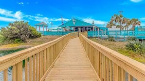 A wooden boardwalk leading to a building with a green roof, surrounded by palm trees and a clear blue sky. - Retreat by the Sea (Myrtle Beach)