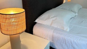 Uma cama com roupa de cama branca, cabeceira escura, mesa de cabeceira com abajur e luminária na parede.