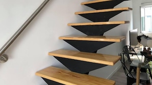 Escadaria moderna com degraus de madeira e faixas verticais pretas, que leva a um espaço com uma mesa de vidro e cadeiras.