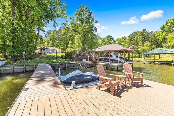 Un muelle de madera con sillas y una tienda de campaña, un barco pequeño, y una casa al fondo.