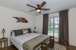 Un dormitorio con cama, ventilador de techo, mesita de noche, lámpara, ventana con cortinas y vista al exterior.